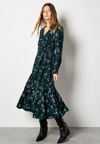 Langärmliges, schwarzes Blumenkleid mit grünen und weißen Blattmustern, gestuftem Saum, tailliertem Schnitt und kombiniert mit schwarzen Absatzstiefeln.