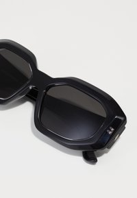 Occhiali da sole oversized neri con montatura geometrica, lenti oscurate e una finitura plastica liscia e lucida. I dettagli notevoli includono particolari metallici.