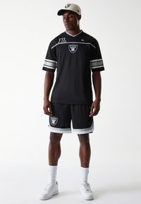 Black athletic jersey with silver stripes and Raiders logo, paired with black shorts featuring logo and white trim, complemented by a beige cap.