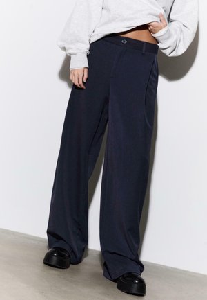 Pantalon à jambes larges en tissu bleu marine foncé, équipé d'une fermeture à boutons, de poches latérales et d'une texture lisse. Associé à des chaussures plateformes noires.