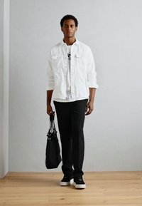 Giacca di denim bianca su una maglietta bianca, pantaloni neri e scarpe nere con suola bianca. Tiene in mano una borsa nera. Pavimento in legno, sfondo chiaro.