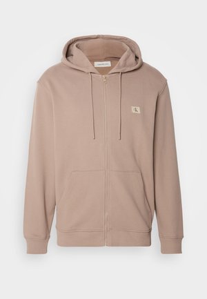 Sudadera con cremallera de color marrón claro, hecha de una tela suave, que presenta una capucha ajustable con cordón, bolsillos frontales, puños acanalados y un pequeño logo en el pecho.