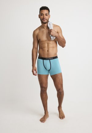 Men's Jockey boxer briefs in light blue cotton with a black elastic waistband, featuring a fitted design and flat seams. A gray towel draped over one shoulder.