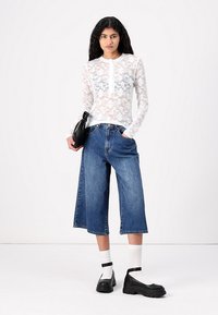 Top alb din dantelă florală cu mâneci lungi, cu o deschidere cu nasturi, asortat cu culottes din denim albastru cu picioare largi și pantofi negri masivi. Șosetele albe sunt vizibile.