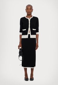 Čierny cardigan s krémovými detailmi a vreckami, skombinovaný s čiernou midi sukňou. Model drží malú štruktúrovanú čiernu kabelku a má na sebe ploché topánky.