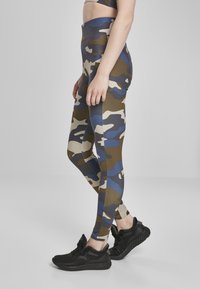 Camo leggings feature a mix of green, blue, and beige patterns, made from a stretchy, fitted material, paired with black athletic shoes.