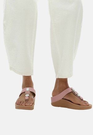 Voeten met roze sleehakken met decoratieve steentjes, gecombineerd met gebroken-witte cropped broek tegen een witte achtergrond.