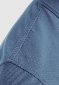 Modrá bavlnená tkanina s textúrovaným povrchom a šitými detailmi, s výrazným švom na pleci. Obsahuje aj dodatočný detail v podobe vrecka.