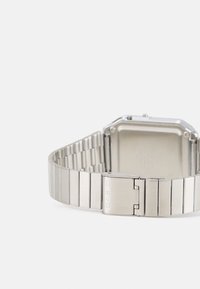 Relógio de pulso metálico com uma caixa quadrada prateada e uma bracelete de aço inoxidável. A bracelete apresenta elos retangulares e um fecho com um logótipo.