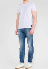 Mies, który ma na sobie białą koszulkę V-neck, niebieskie dopasowane dżinsy i białe sneakersy, stojący na tle jednolitego jasnego tła.