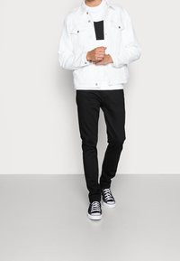 Giacca di denim bianca con bottoni argentati, t-shirt nera con un motivo orizzontale, pantaloni neri slim-fit e sneakers nere con suola bianca.