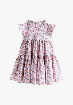 Junges Mädchen mit Zöpfen, das ein Blumenmuster-Kleid und gelbe Socken trägt, steht vor einem Hintergrund mit einem Blumenmarkt aus Pappe.