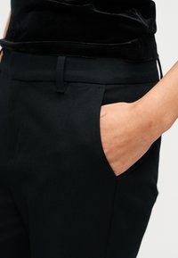 Main insérée dans la poche latérale d'un pantalon noir sur mesure porté avec un haut en velours noir.