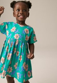 Lyhythihainen mekko vihreästä kankaasta, jossa on vaaleanpunaisia ja sinisiä kukkakuoseja, rypytetty hame ja pyöreä pääntie.
