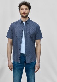 Korte mouwen, knoopshirt in donkergrijs met een subtiele zeilbootprint, gecombineerd met een lichtblauw T-shirt en blauwe jeans.