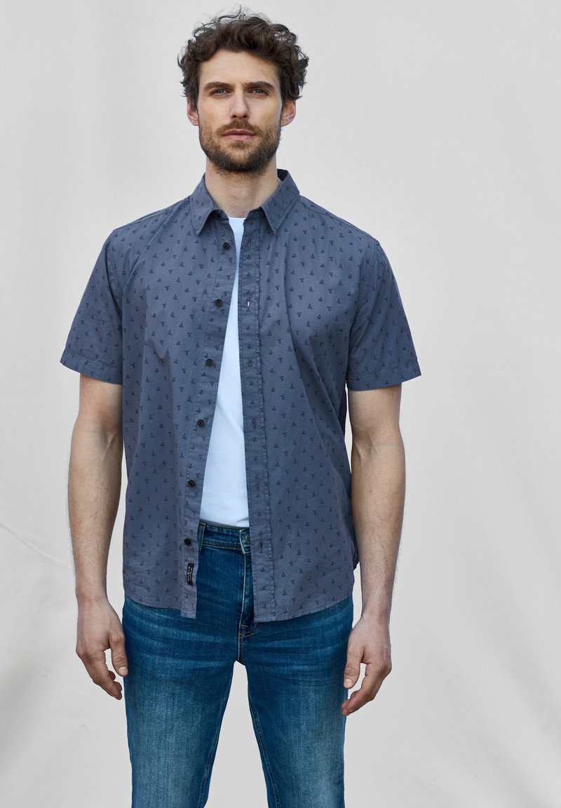 Korte mouwen, knoopshirt in donkergrijs met een subtiele zeilbootprint, gecombineerd met een lichtblauw T-shirt en blauwe jeans.