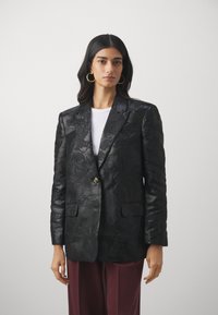 Svart blazer med blomstermønster, enkel knappelukking, slagkrage og to frontlommer. Brukt over en hvit topp og burgunderbukser.