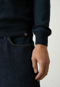 Dunkelblauer Pullover aus glattem Material, mit gerippten Bündchen und Saum, versehen mit einem kleinen "Colmar"-Logotag an der Seite, kombiniert mit dunkler Denim-Jeans.
