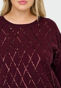 Burgundowy sweter z teksturowanym splotem, który ma wzór w diamenty wykonany z czarnych i różowych cekinów, okrągły dekolt i luźny krój.