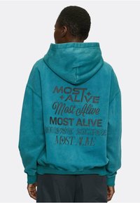 Felpa oversize color teal con grafiche testuali varie in nero, design a pullover e cappuccio spazioso. Tessuto morbido e strutturato.