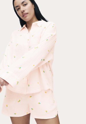 Vrouw in een lichtroze pyjamaset met kleine gele en groene bloemenprints, staand tegen een effen lichte achtergrond.