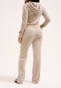 Beige fluwelen tweedelig set met een rhinestone "Juicy"-logo op de achterkant van de hoodie en de joggingsbroek. Heeft een aansluitend ontwerp en een kap.