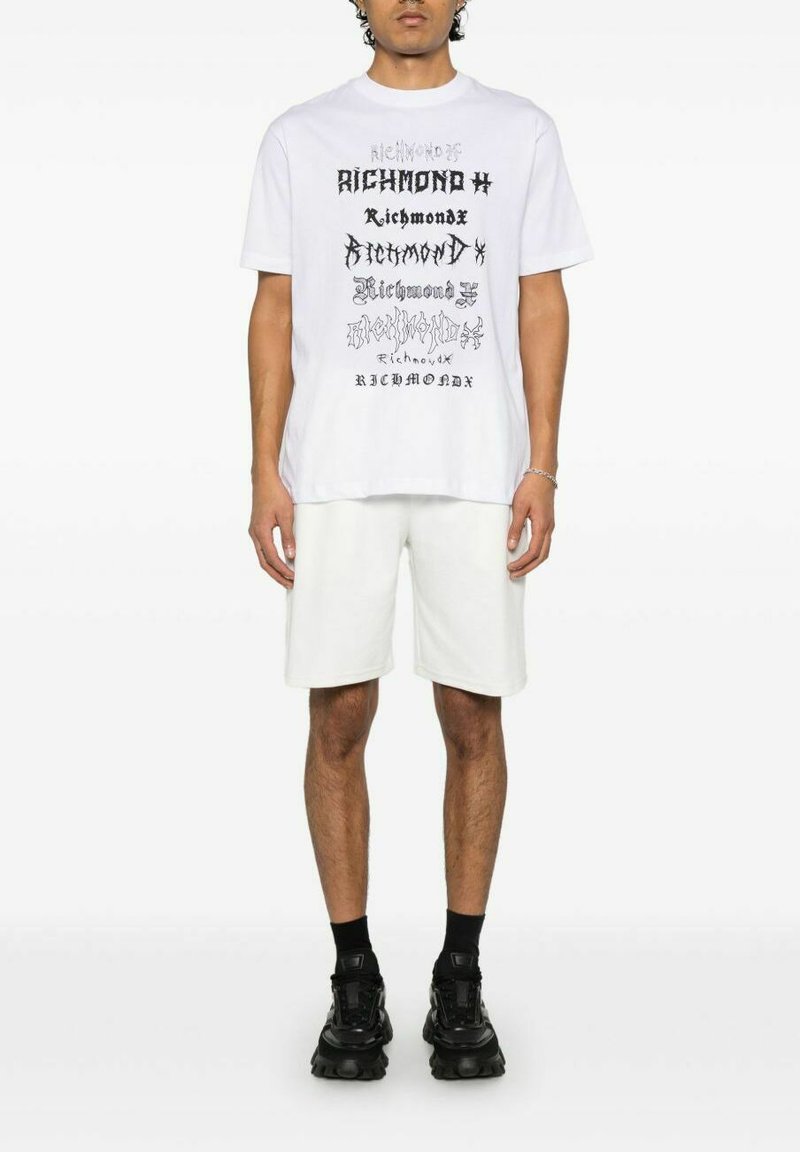 T-shirt bianca con testo grafico nero in vari caratteri, abbinata a pantaloni corti bianchi e sneakers nere. Design semplice, maniche corte e vestibilità casual.