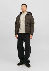 Puffervest i mørkebrun, med hætte og quiltet design. Båret over en lysebrun halv-zip sweatshirt og mørke jeans.