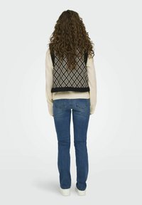 Gebreid vest met ruitpatroon in zwart en crème, gecombineerd met lichtblauwe jeans en witte sneakers, gezien van achter. Krullend haar zichtbaar.