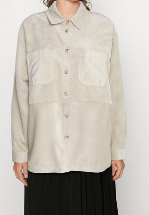 Beige corduroy button-up shirt with two large front pockets worn over a black pleated skirt.