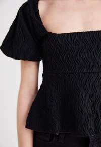 Top nero testurizzato con scollatura quadrata e maniche a sbuffo, caratterizzato da un motivo ondulato e una silhouette a peplo. Abbinate a jeans scuri.