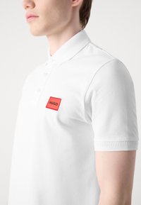 Hvid polo shirt med tekstureret stof, en ribbet krave og en rød rektangulær lapp med "HUGO" i sort skrift på venstre side.