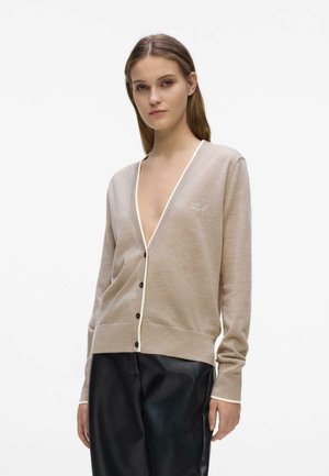 Young woman wearing beige buttoned cardigan with white trim and black leather pants, standing against plain light background.