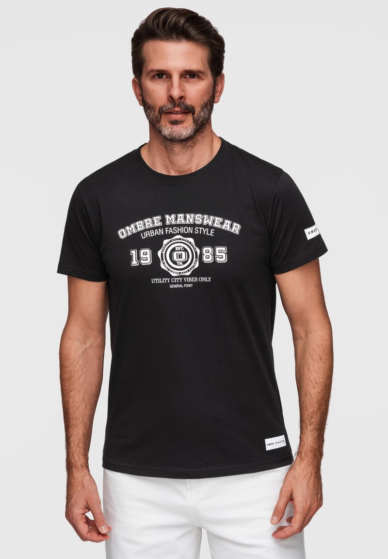 Homme aux cheveux courts et foncés avec une barbe, portant un t-shirt noir avec un texte blanc et un pantalon blanc, debout devant un fond clair.