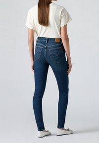 Jeans ajustados de mezclilla azul con cintura alta, bolsillos traseros con costura curva y una etiqueta de cuero marrón. Combinados con una camiseta blanca y zapatos planos.