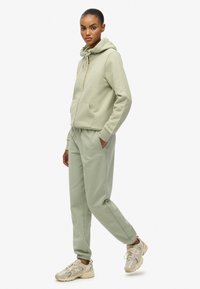 Jasnozielona bluza z kapturem na zamek, z przednią kieszenią i sznurkiem. W połączeniu z dopasowanymi, zwężającymi się spodniami dresowymi. Buty sportowe w neutralskich kolorach.