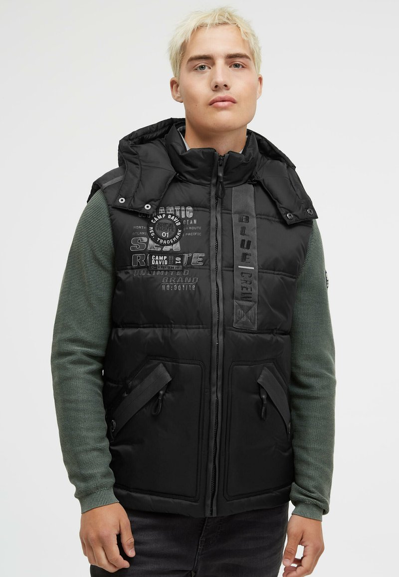 Schwarze gepolsterte Weste mit hohem Kragen, Front-Reißverschluss und zwei seitlichen Taschen. Verfügt über gedruckte Texte und Logo-Akzente auf einer glatten Oberfläche.