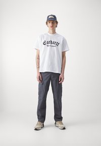Weißes T-Shirt mit kurzen Ärmeln und schwarzem Text, kombiniert mit grauen Cargo-Hosen. Das Model trägt eine blaue Mütze und beige Sneakers. Minimalistischer Hintergrund.