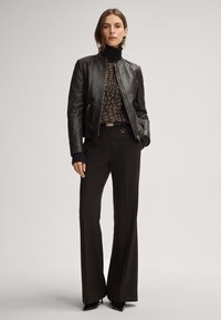Schwarze Lederbomberjacke mit Reißverschluss, geripptem Kragen und Schulterdetails, kombiniert mit braunen Schlaghosen und schwarzen High Heels.