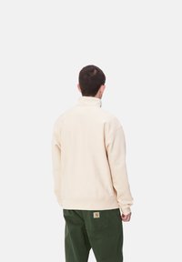 Sudadera de forro polar beige con cuello alto, vista desde la parte posterior, combinada con pantalones verde oscuro. Textura suave y diseño relajado.