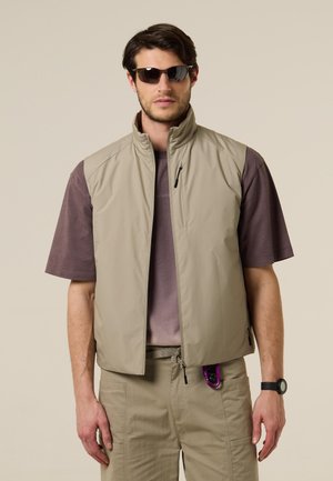 Mann mit Sonnenbrille, beiger ärmelloser Reißverschlussweste, braunem Kurzarmhemd, beiger Hose mit lila Karabiner und schwarzer Armbanduhr.