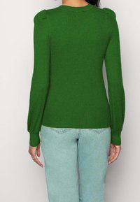 Zielony sweter z prążkowanej dzianiny z bufkami na ramionach, długimi rękawami i dopasowanym krojem, w zestawieniu z jasnoniebieskimi dżinsami.