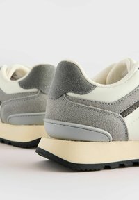 Graue und cremefarbene Sneakers mit einem Obermaterial aus Wildleder und Leder, ausgestattet mit einem gepolsterten Kragen, strukturierten Akzenten und einer kontrastierenden Gummisohle.