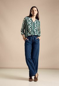 Blusa verde con motivo geometrico e maniche lunghe, abbinata a pantaloni in denim blu larghi e scarpe slip-on marroni.