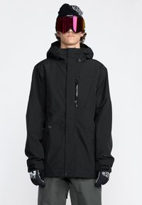 Veste imperméable noire avec capuche, poche zippée sur le devant et poignets élastiques. Le modèle porte des lunettes de protection roses réfléchissantes et des gants noirs avec un design graphique.