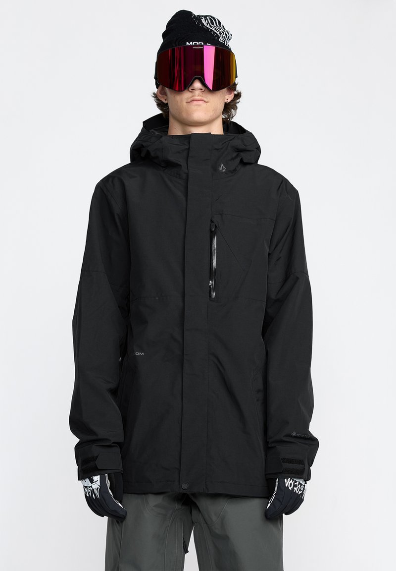 Veste imperméable noire avec capuche, poche zippée sur le devant et poignets élastiques. Le modèle porte des lunettes de protection roses réfléchissantes et des gants noirs avec un design graphique.