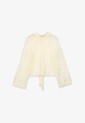 Cape blanc cassé à texture plissée, parsemée de sequins. Comprend un col montant et des liens à la taille.