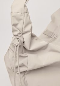 Grand sac fourre-tout beige clair en tissu texturé, avec une anse froncée et des fermetures à cordon élastique pour un accès sécurisé.