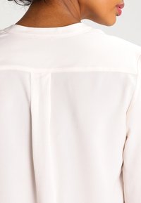 Lehká, bledá blůza s hladkým povrchem, s jednoduchým zadním švem a kulatým výstřihem. Měkký, jemný design látky.
