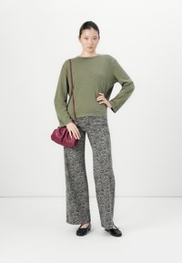 Donna in piedi con una mano su un fianco, che indossa un maglione verde oliva, pantaloni grigi a stampa zebra, ballerine nere e tiene in mano una piccola borsa burgundy.